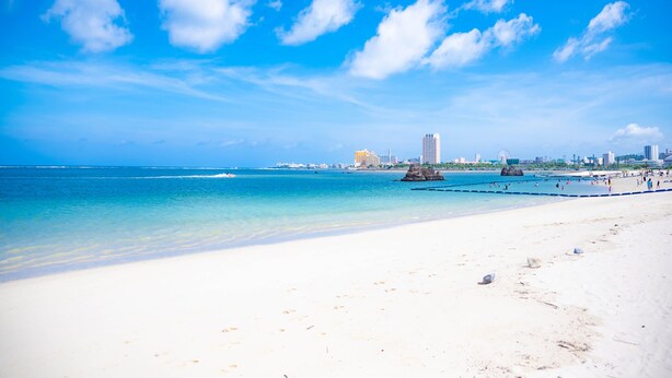 Una playa de arena blanca, agua azul cristalina y un horizonte urbano al fondo.