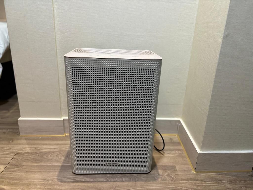 Un altavoz gris rectangular con panel frontal perforado, sobre un suelo de madera frente a una pared con detalles amarillos.