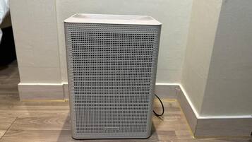 Un altavoz gris rectangular con panel frontal perforado, sobre un suelo de madera frente a una pared con detalles amarillos.