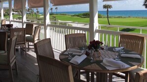 A covered outdoor dining area with tables and chairs, overlooking a golf course and the ocean.