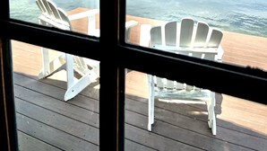 A view through a window of a wooden deck with white Adirondack chairs and a table, overlooking a calm lake with trees and a clear sky.