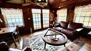 A cozy cabin interior with a wooden ceiling, a leather sofa, a dining table with chairs, and a large window offering a view of a lake.