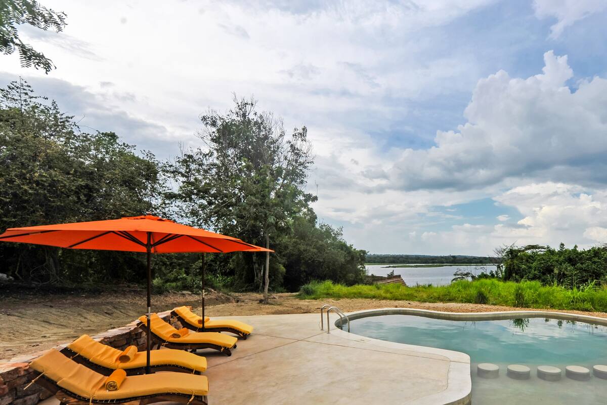 A poolside area with lounge chairs and an umbrella, surrounded by greenery and a view of a lake.