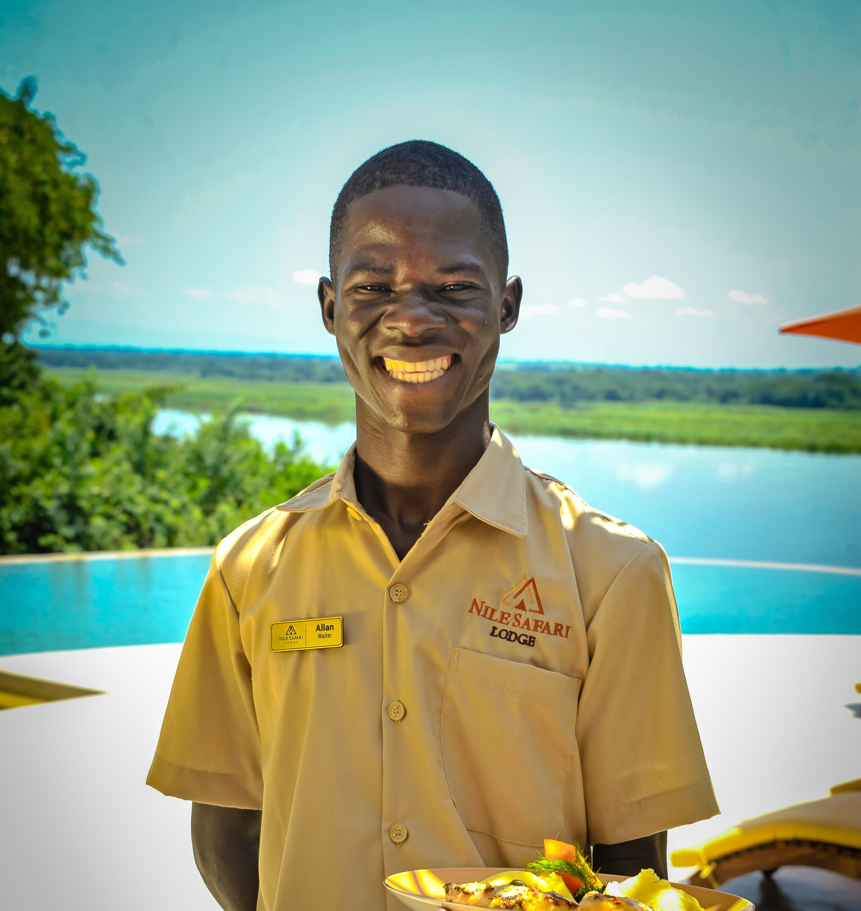 A person in a yellow shirt with a name tag and lodge logo, holding a plate of food, with a pool and greenery in the background.