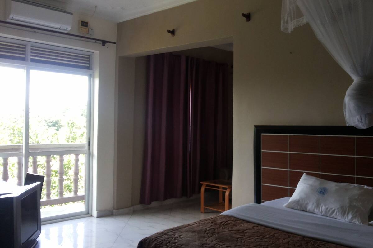 A hotel room with a bed, a small table, a chair, a window with curtains, and a balcony.