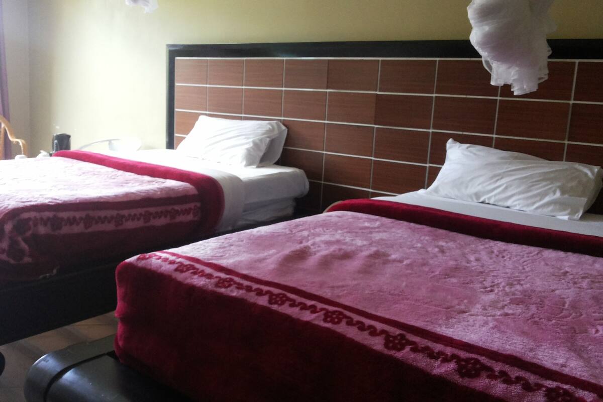 Two beds with red and white bedding, a wooden headboard, and a chair in a room with a tiled floor.