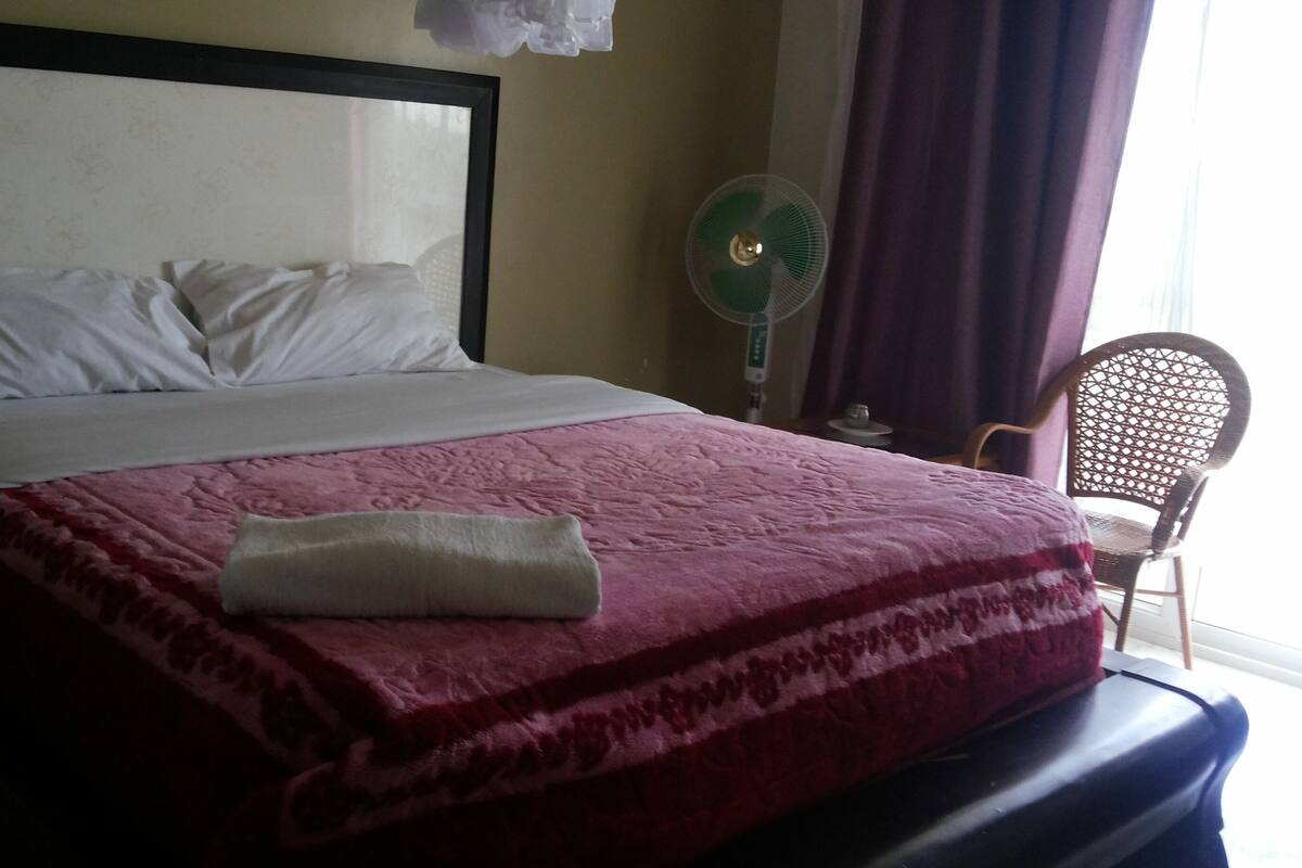 A neatly made bed with white pillows and a pink blanket, a standing fan, and a chair by the window.