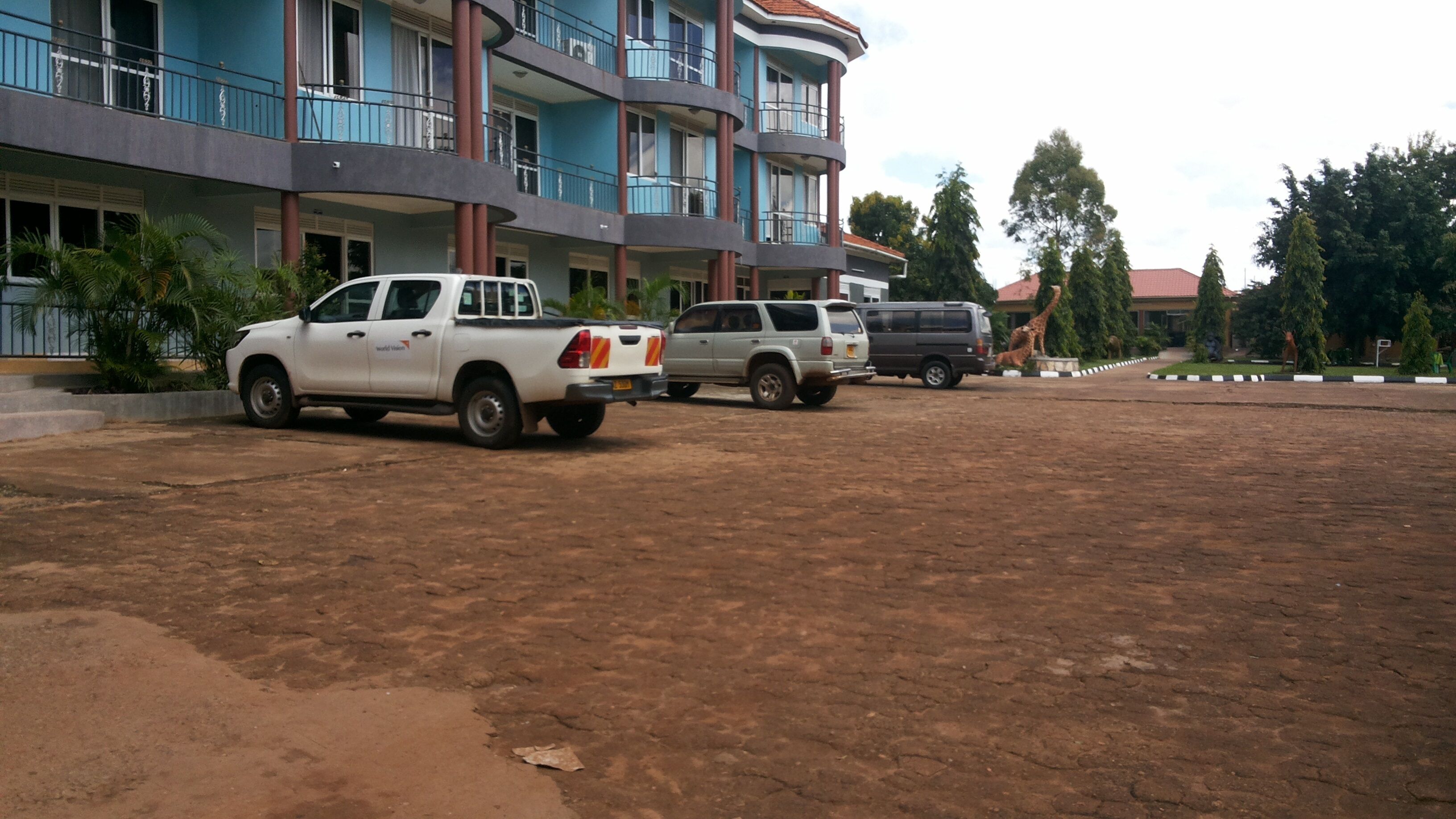 A parking area with several vehicles, including a white pickup truck and a grey SUV. In the background, there is a multi-story building with balconies and a red roof.
