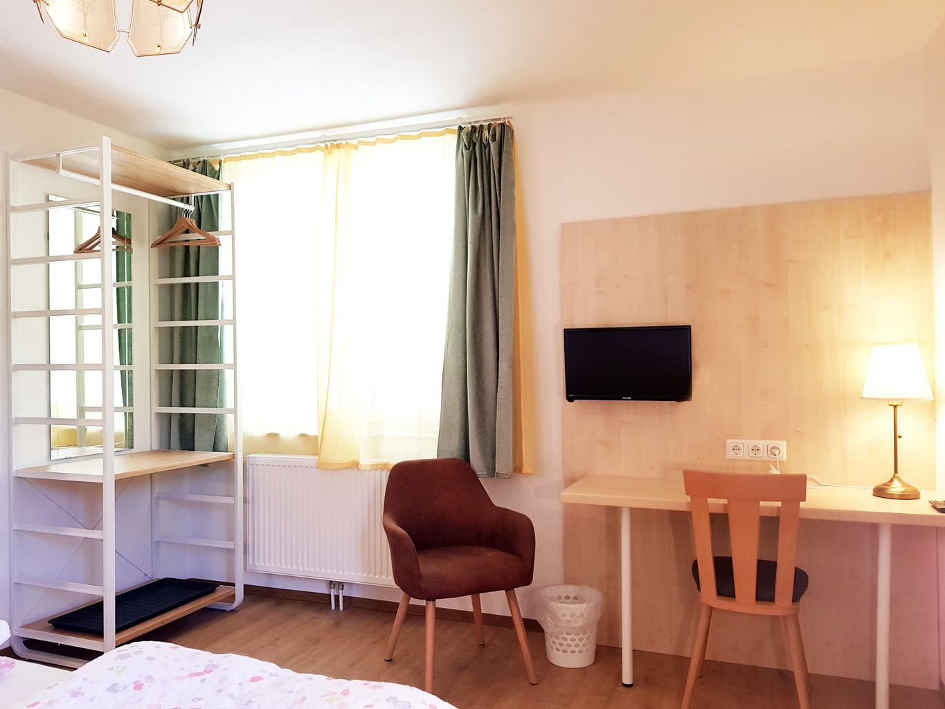 A hotel room with a bed, a desk with a chair, a TV, a wardrobe, and a radiator.