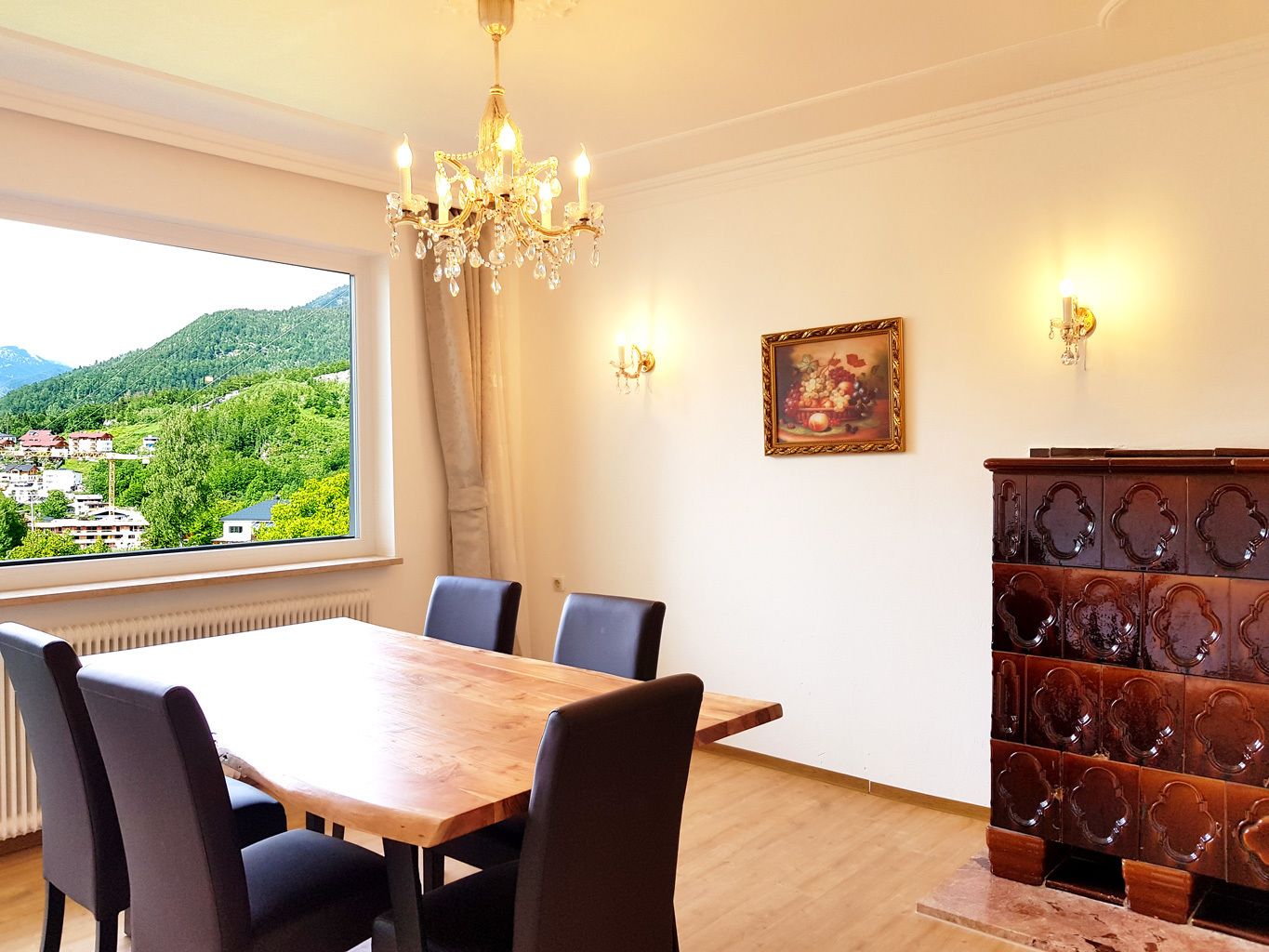 A dining room with a wooden table and chairs, a chandelier, a painting, and a view of a green landscape through a window.