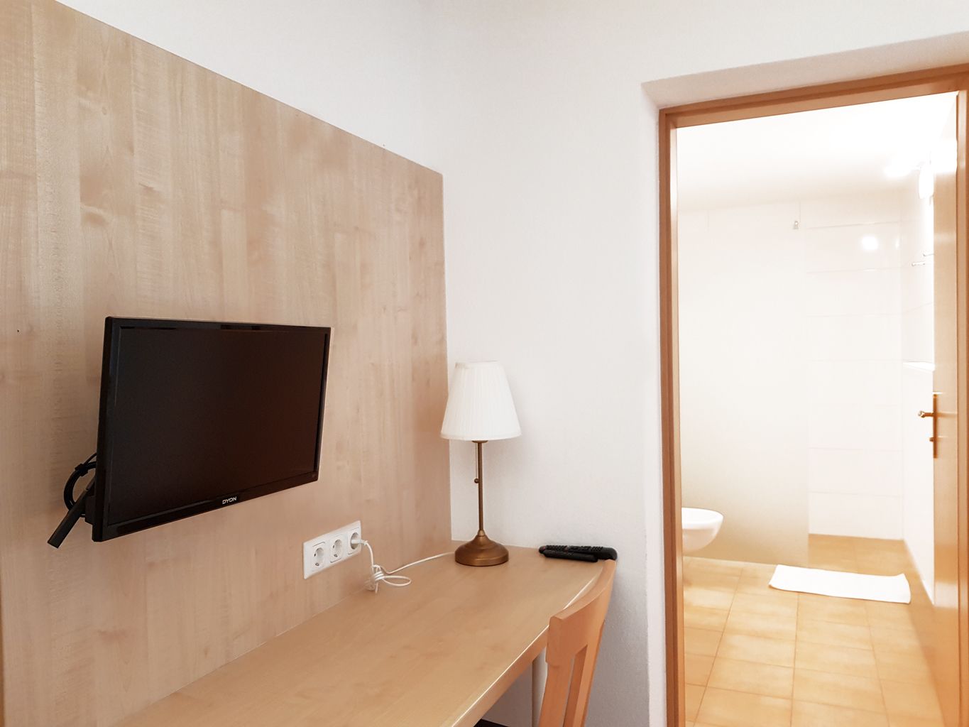 Una habitación con un escritorio de madera, un televisor montado, una lámpara y un control remoto.