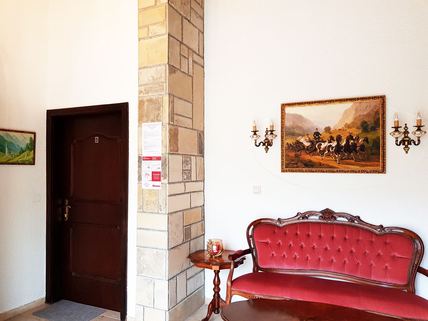 Una habitación con puerta de madera, un banco tapizado en rojo, una mesita con una vela y un cuadro en la pared.