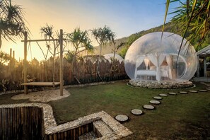 Una estructura de cúpula transparente con una cama en su interior, rodeada por un camino de piedra, valla de bambú y palmeras.