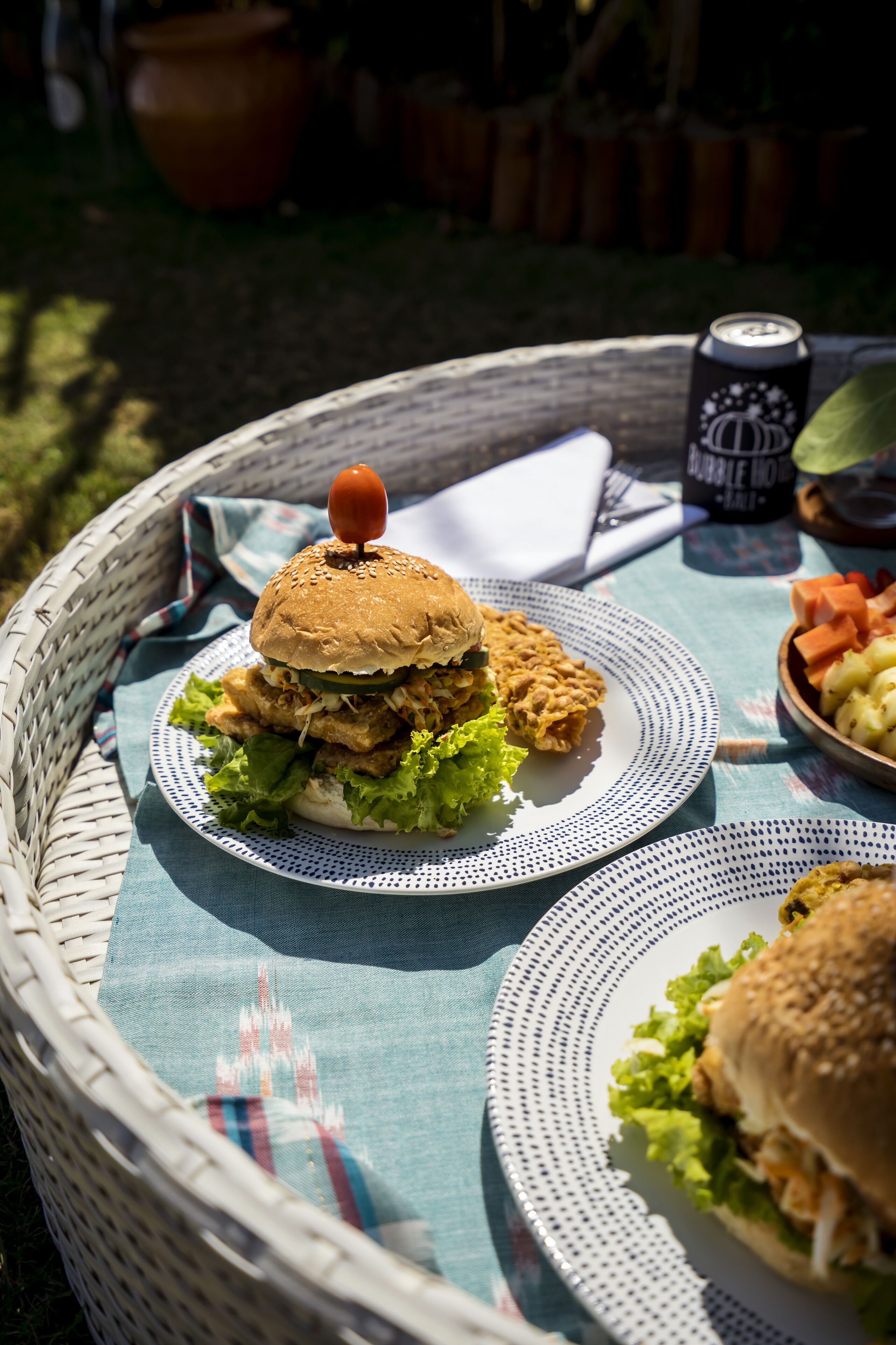 A wicker basket containing a burger with a tomato on top, a can of Double MOCHA, and a bowl of vegetables on a table with a blue cloth.