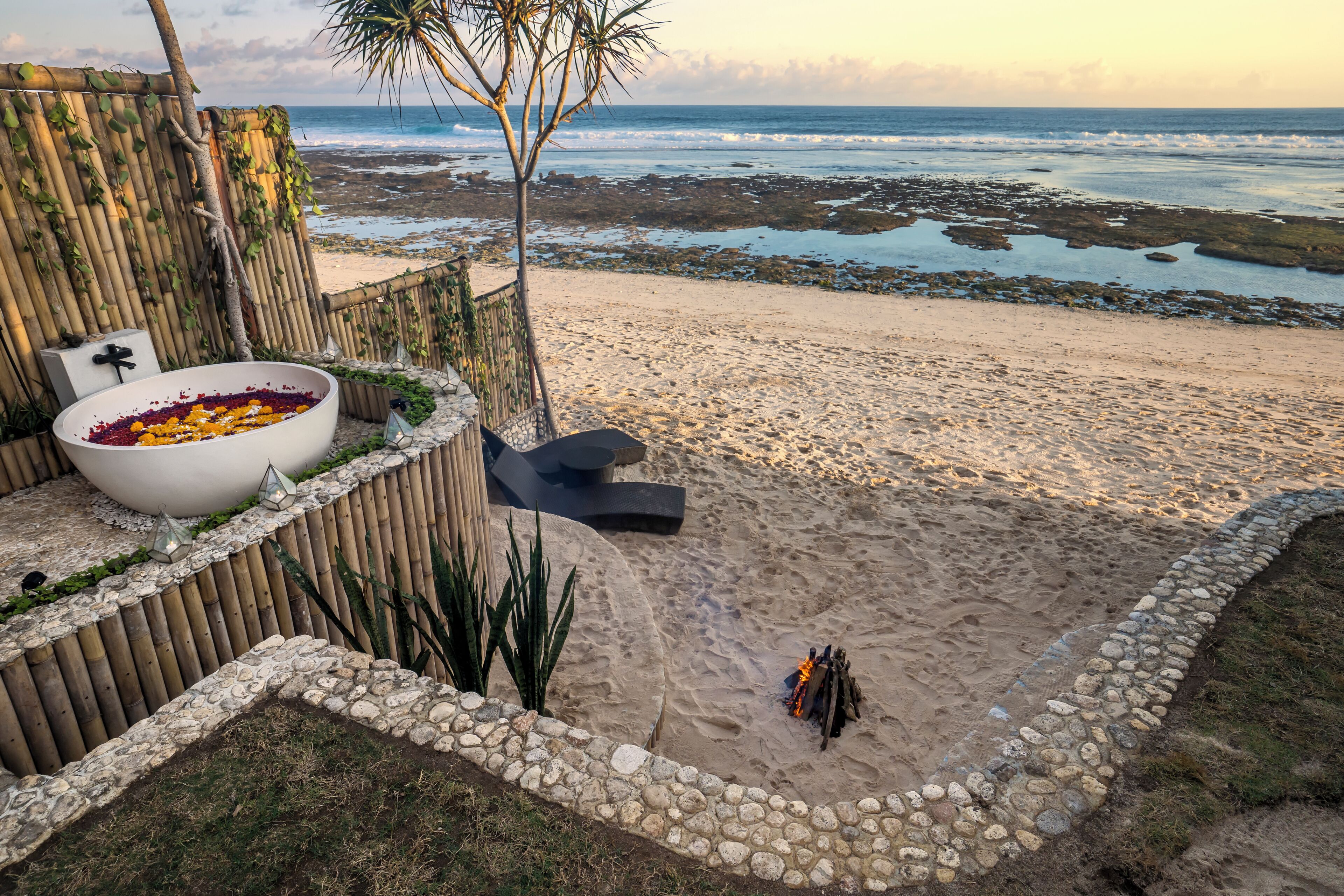 Une terrasse au bord de la plage, avec un mur en pierre, un grand vase blanc rempli de fleurs et une vue sur l’océan.