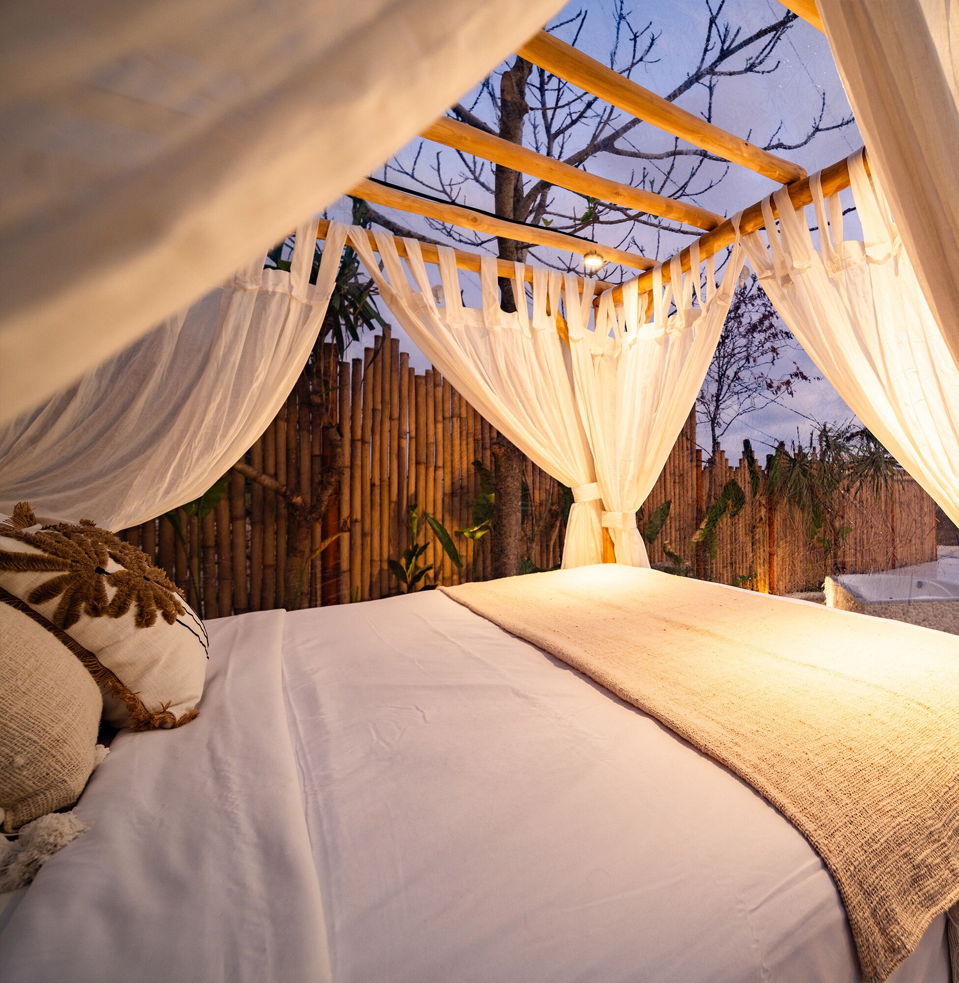 A bed with white linens and a canopy, a wooden trellis, and a bamboo fence in the background.