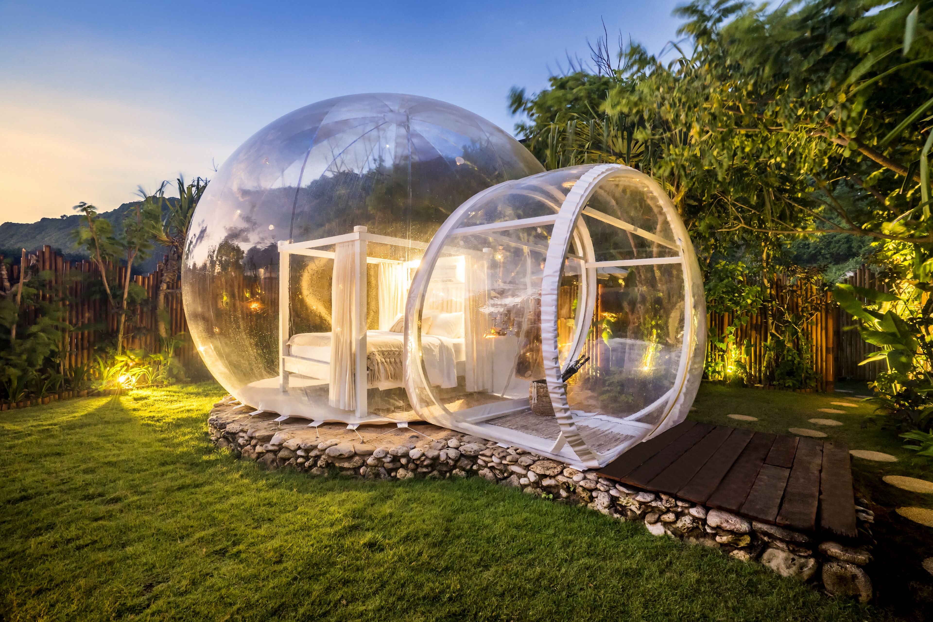 A transparent dome structure with a bed and seating area, surrounded by a stone base and lush greenery.