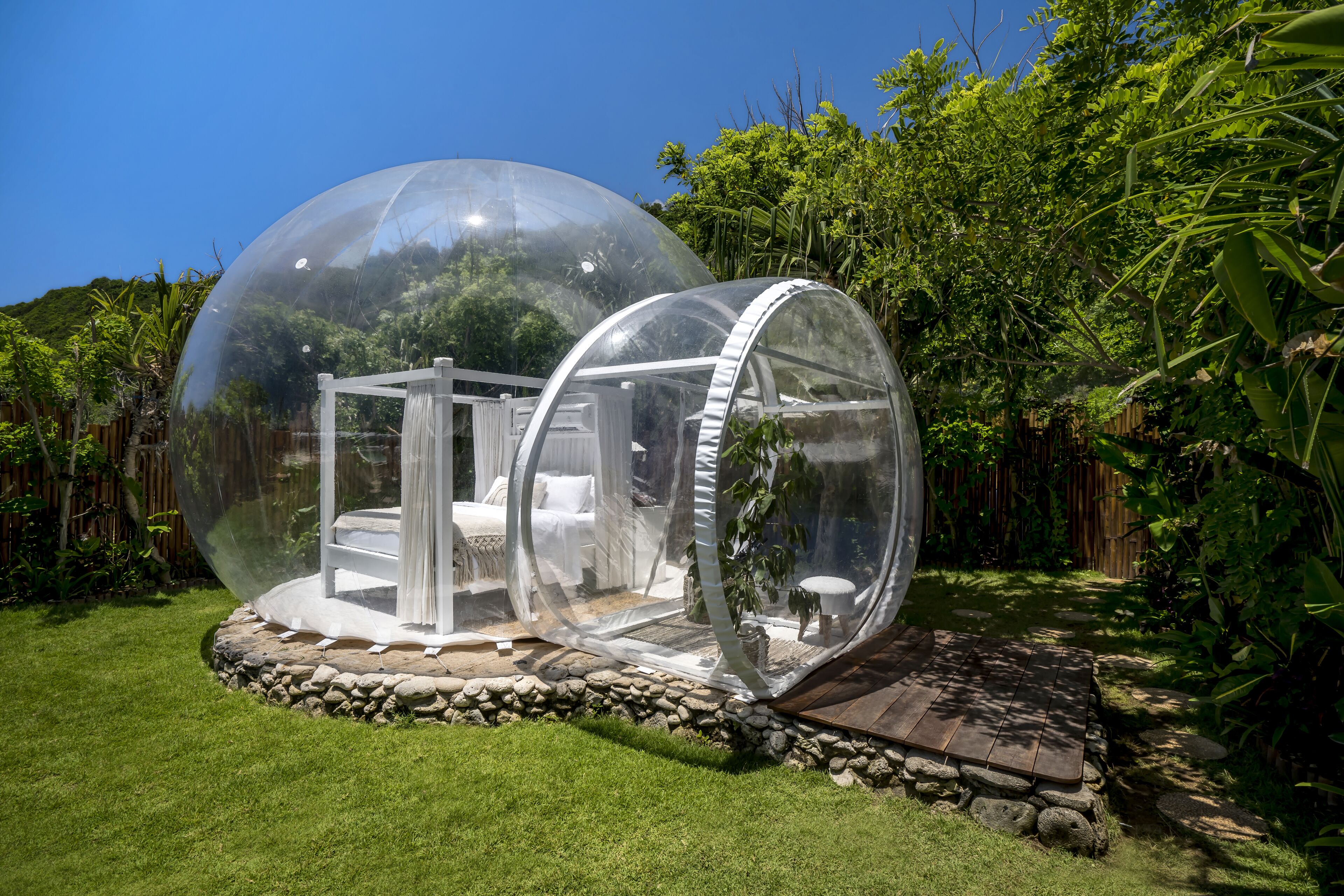Dos estructuras esféricas transparentes con una cama y zona de estar en su interior, rodeadas por una base de piedra y abundante vegetación.