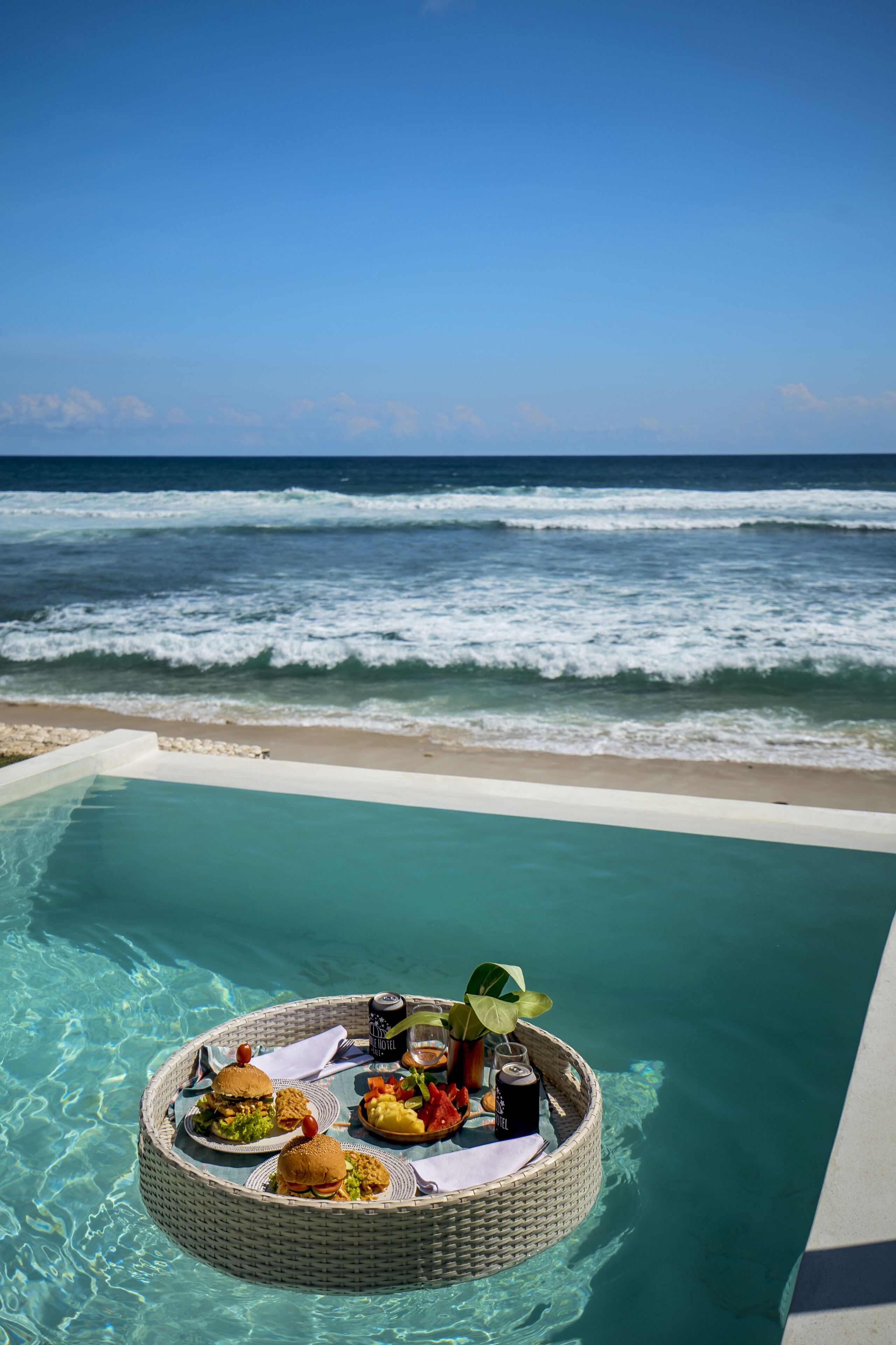 A poolside dining setup with a wicker table floating in clear water, surrounded by a beach and ocean view.