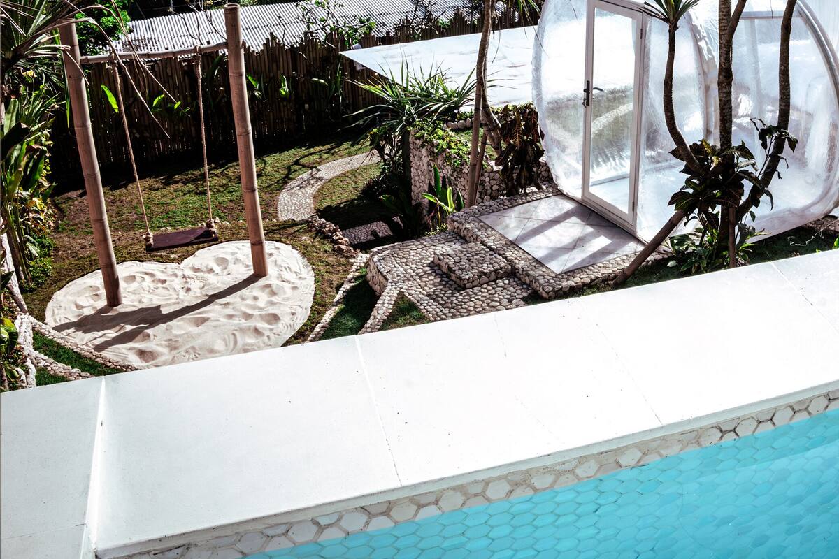 Une piscine avec un bord carrelé, une balançoire et un espace jardin agrémenté de plantes et d’une structure en bois.
