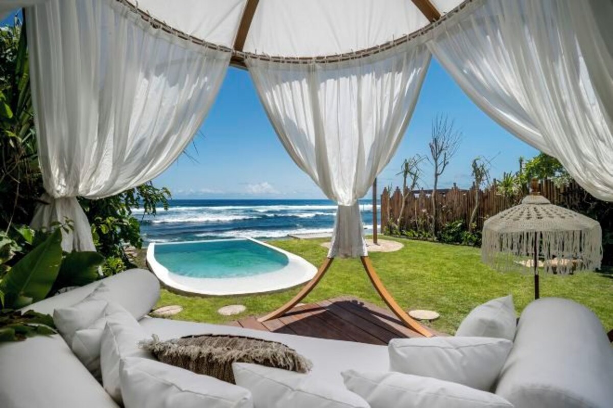 A beachside lounge with a view of the ocean, a pool, and a thatched umbrella.