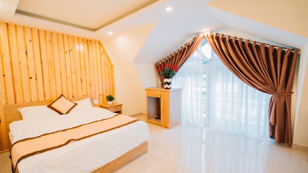 Un dormitorio con cabecera de madera, cama con ropa de cama blanca, mesita de noche con una planta y una ventana con cortinas.