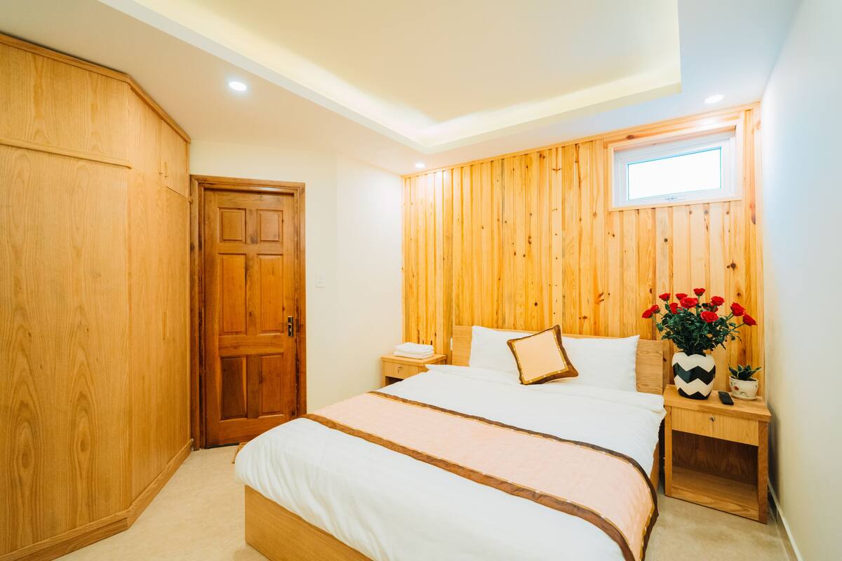 Una camera da letto con un'armadiatura in legno, un letto con biancheria bianca, un comodino con un vaso di fiori rossi e una finestra con vista sull'esterno.