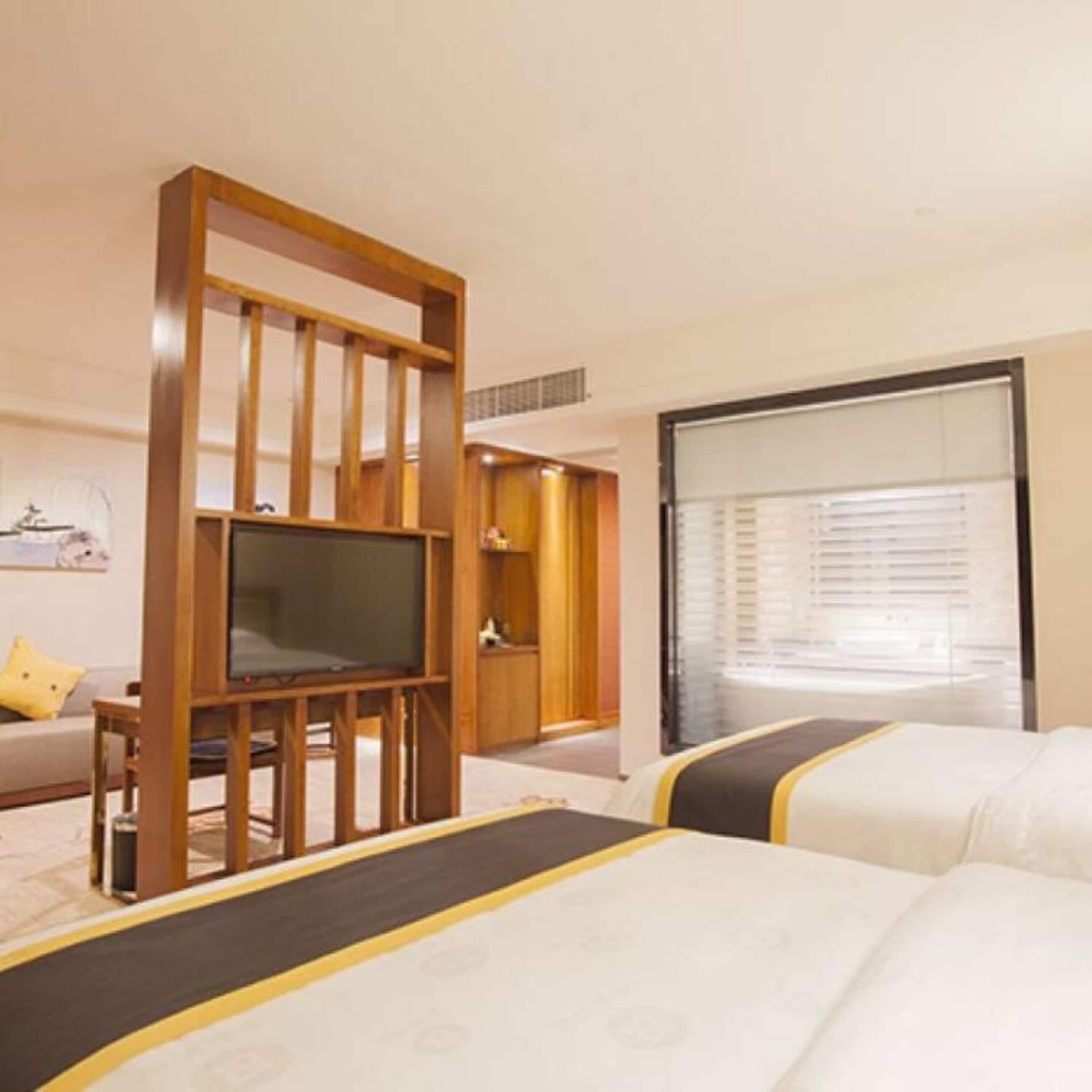 A hotel room with a large bed, a TV on a wooden stand, a sofa, and a window with blinds.