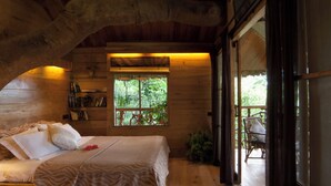 A wooden cabin room with a bed, a nightstand with a lamp, a window with a view of greenery, and a large tree branch arching over the room. - Tranquil Resort - BluSalzz Collection, Wayanad (Sulthan Bathery)