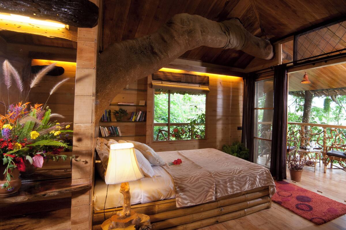 Un dormitorio rústico de madera con una cama, una lámpara de noche, un cabecero de madera y una ventana con vistas a la vegetación.