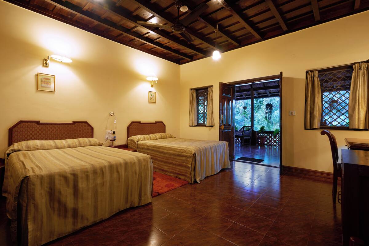 Habitación de hotel con dos camas, vigas de madera en el techo, suelo de baldosas y vistas a un espacio exterior con plantas a través de una puerta abierta.