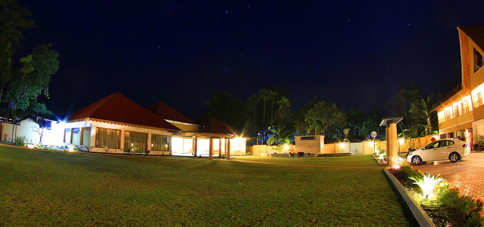 A well-lit residential area at night with multiple houses, a paved driveway, and a car parked on the right.