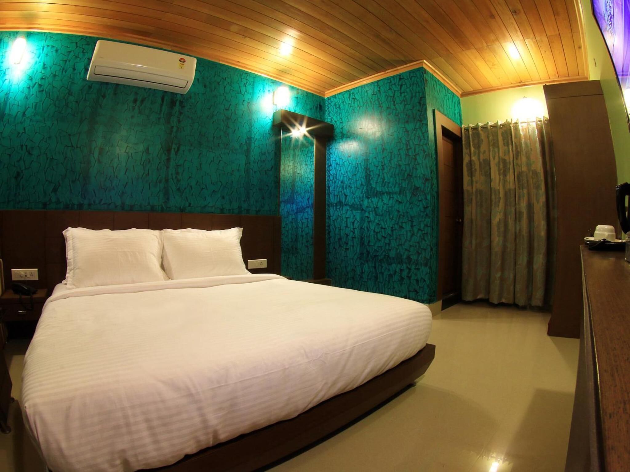 Een hotelkamer met een groot bed, houten plafond, teal muren met structuur, een airconditioningapparaat, een nachtkastje, een bureau met telefoon en een gordijn.