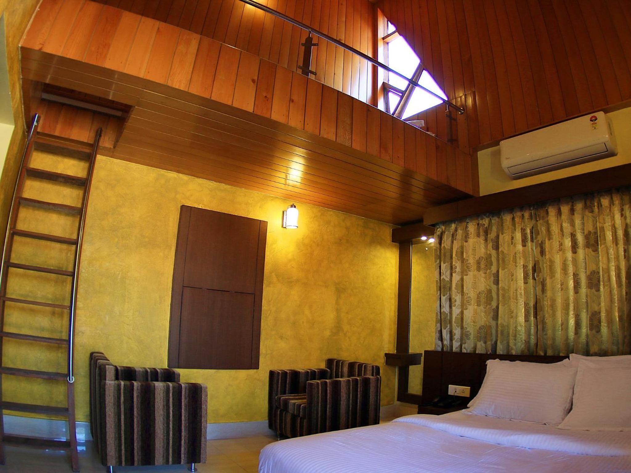 A hotel room with a wooden ceiling, a bed, a desk, a chair, a ladder, and a window.