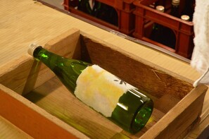 A green glass bottle with a white label in a wooden crate. - Satoyama Guest house COUTURE - Hostel (Ayabe)