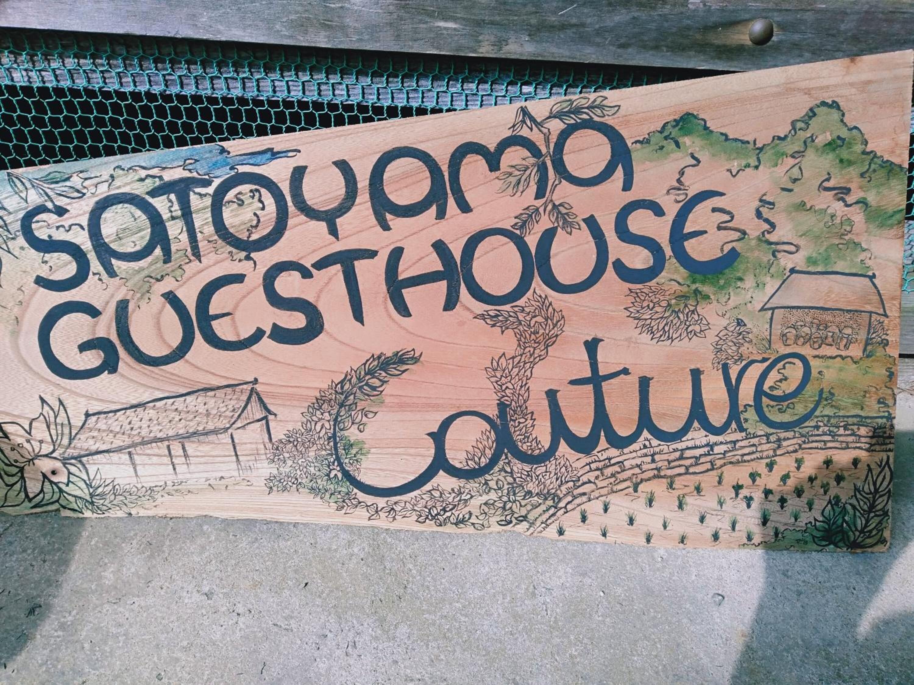 A wooden sign with the text "Sato-yama Guesthouse Culture" and illustrations of a house, trees, and a landscape.