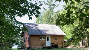 A small wooden cabin with a front porch, surrounded by trees and a grassy yard.