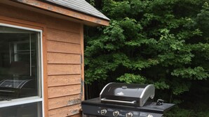 A wooden deck with a barbecue grill and a window overlooking a backyard with trees.