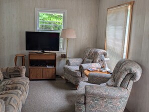 Una sala de estar con un televisor de pantalla plana, un mueble de madera para entretenimiento, sillones estampados, una lámpara de pie y una ventana con persianas.