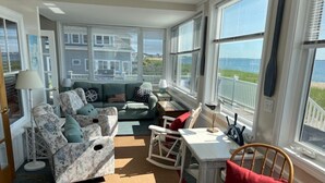 A sunlit room with a view of the ocean, featuring a sofa set, a dining table, and a rocking chair.