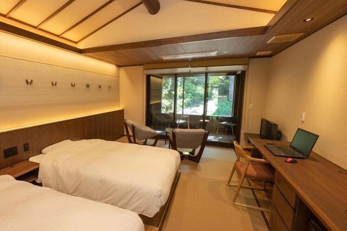 A hotel room with two beds, a desk with a laptop, and a view of the outdoors.