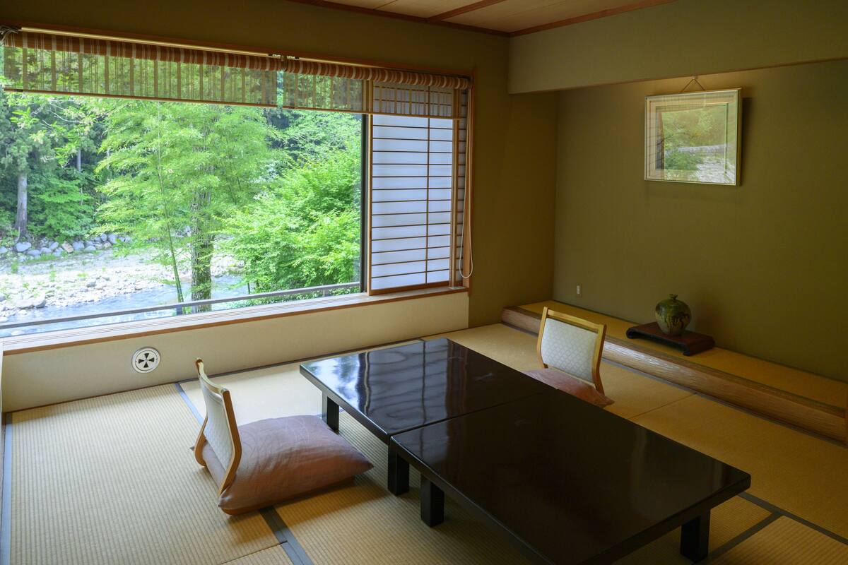 A tatami mat room with a low table and chairs, a window with shoji screens, and a framed picture on the wall.