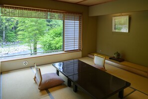 A tatami mat room with a low table and chairs, a window with shoji screens, and a framed picture on the wall.
