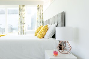 A neatly made bed with white and yellow patterned pillows, a grey upholstered headboard, and a bedside table with a white lamp and a pink candle.