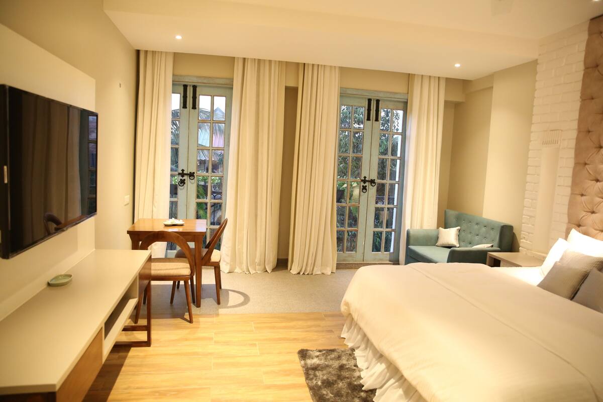 Un dormitorio con cama, escritorio, sillón, sofá y ventanas amplias con cortinas.