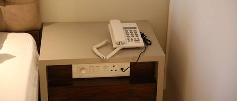 A hotel room with a bed, a bedside table with a telephone, and a power outlet.