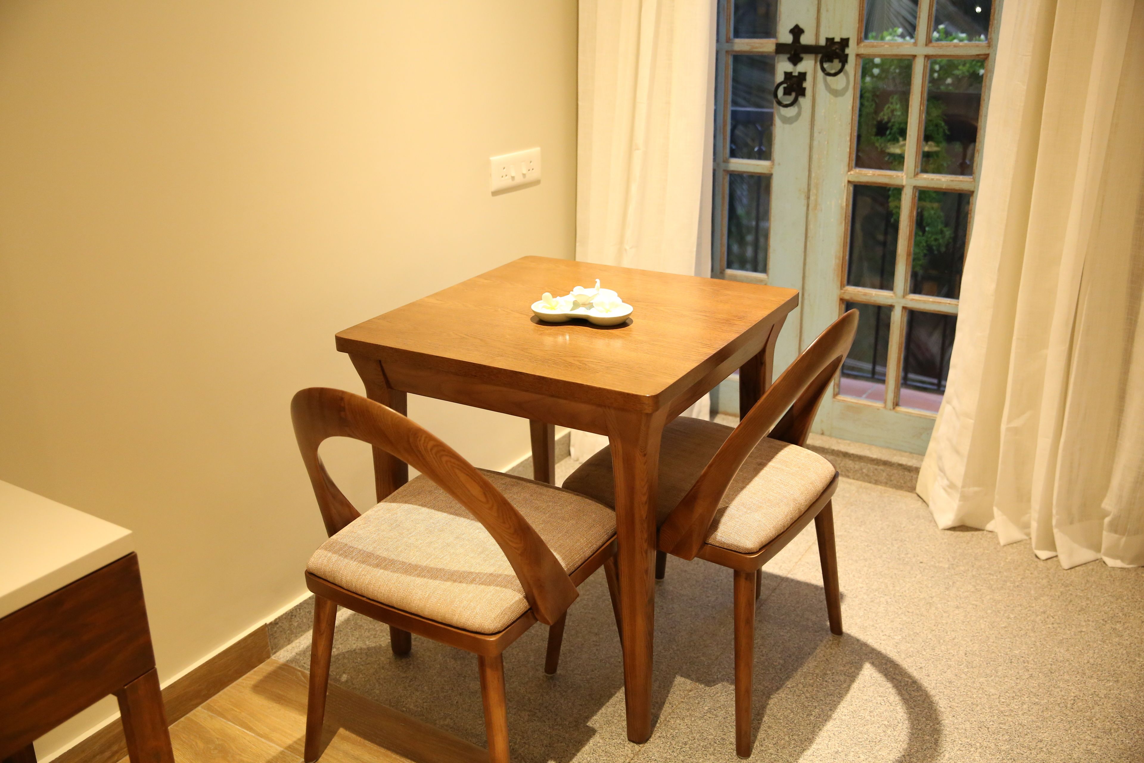 A dining area with a wooden table and four chairs, a small white plate on the table, and a window with curtains.