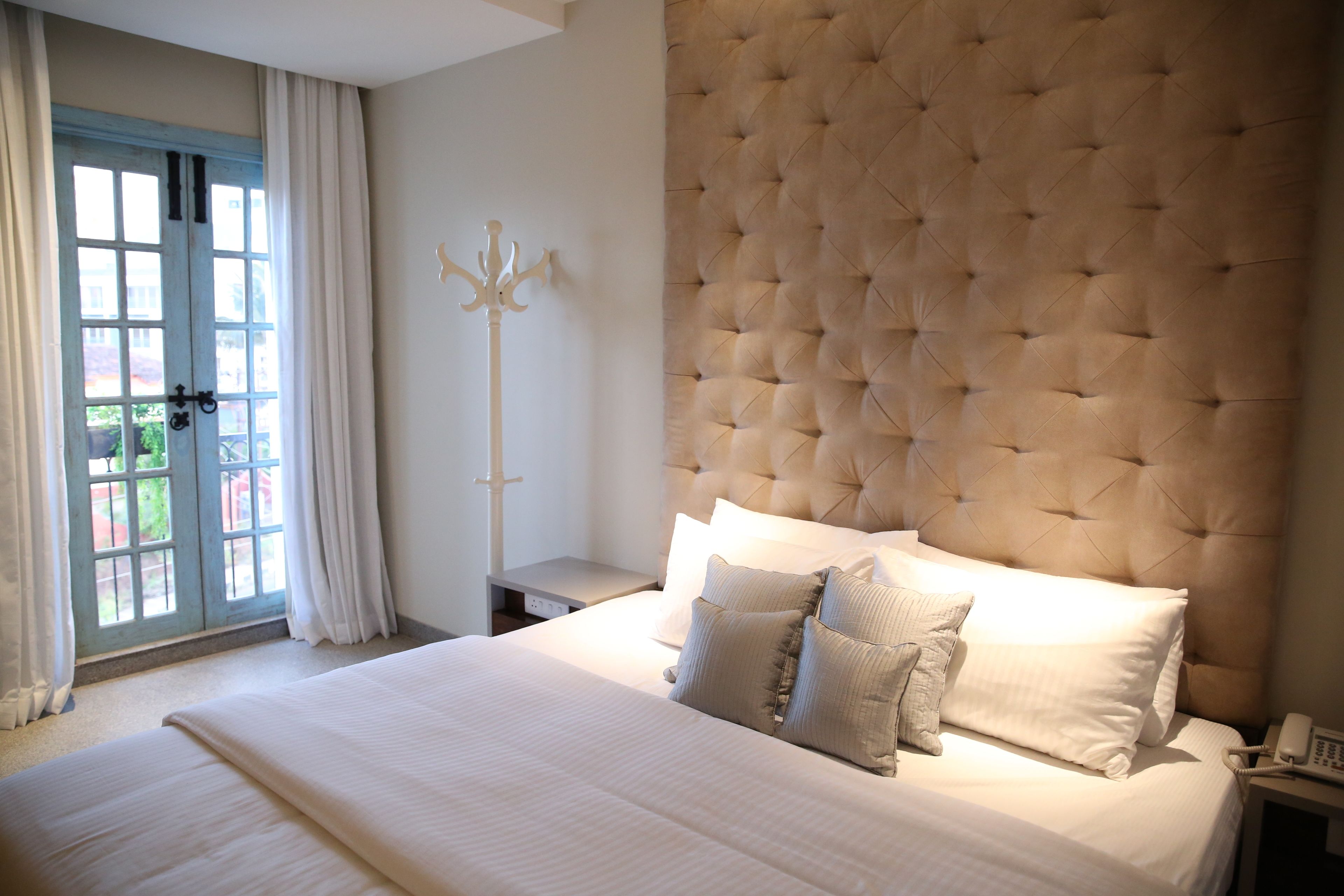 Un lit bien fait, avec du linge de lit blanc et plusieurs oreillers, une tête de lit capitonnée, une table de chevet avec une lampe et un porte-manteau.