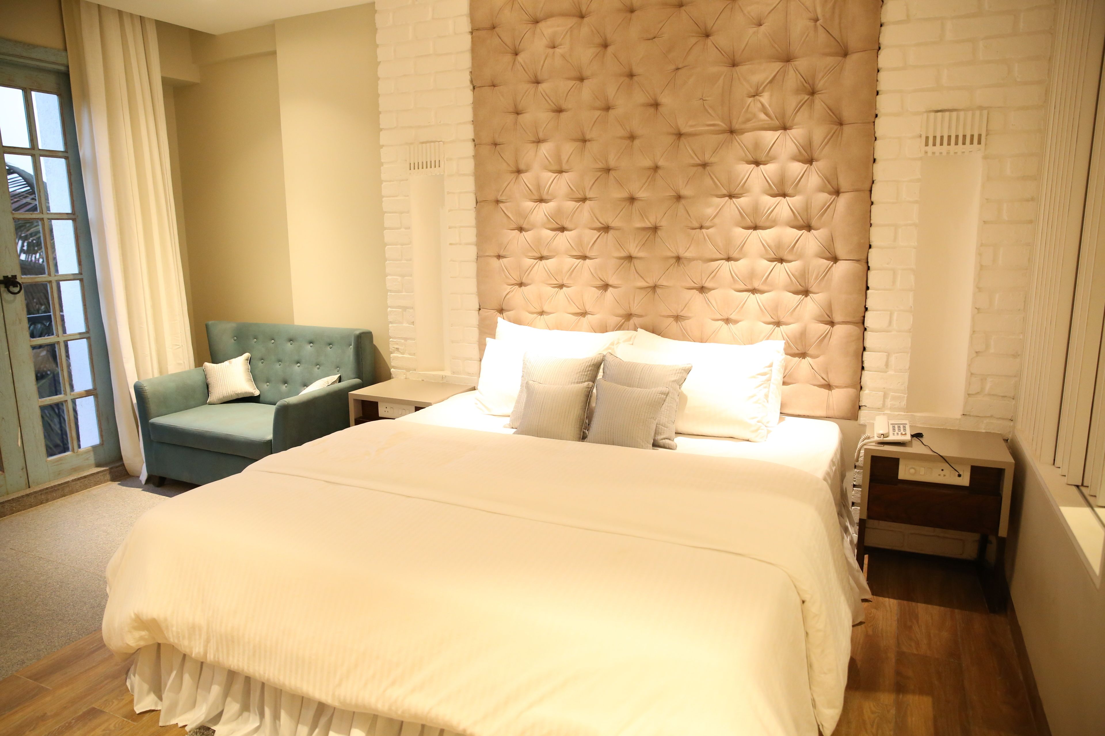 Una cama grande con cabecera capitonada, un sillón verde y una mesita de noche con una lámpara.
