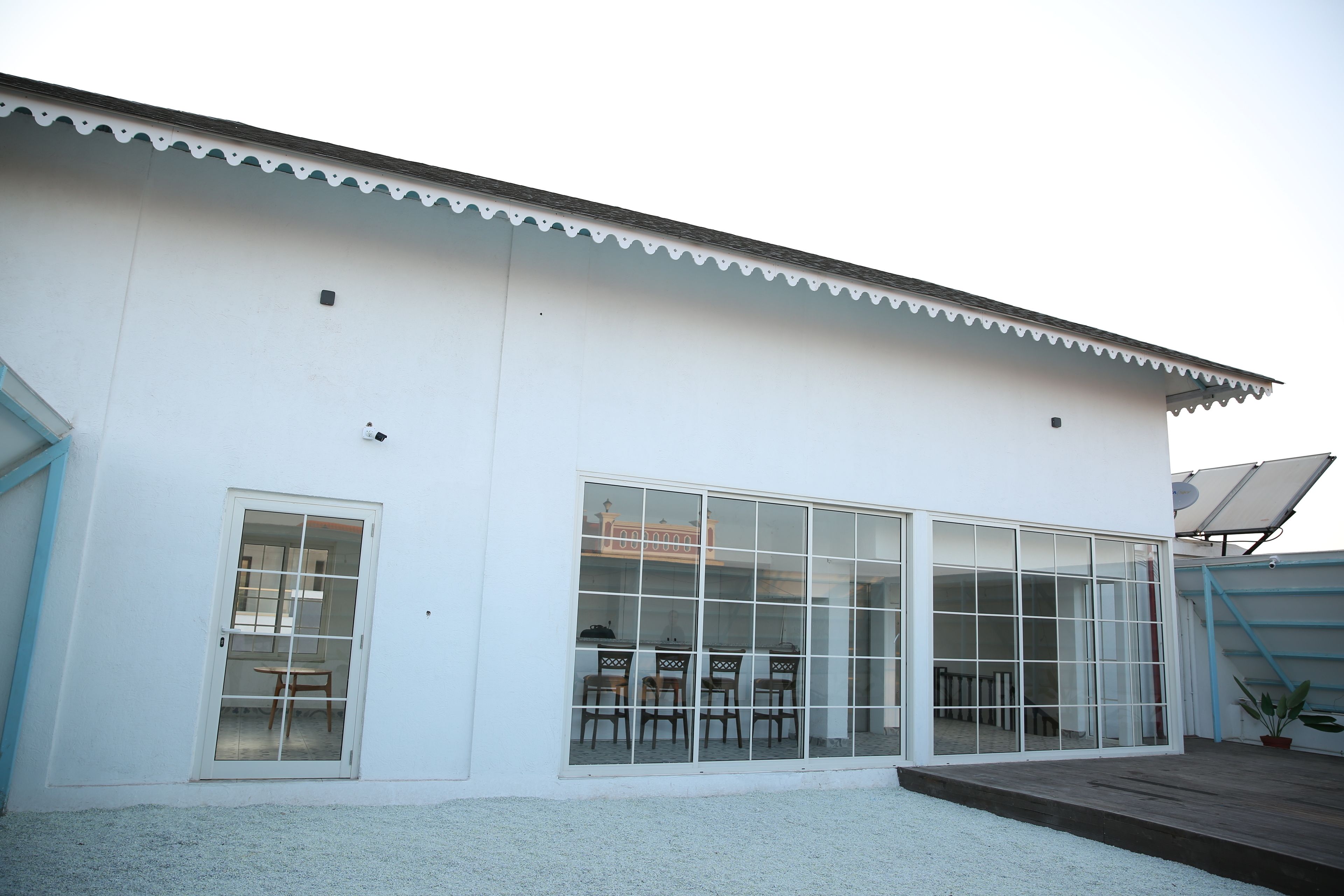 A white building with large windows and a door, a small outdoor area with a table and chairs, and a solar panel on the roof.